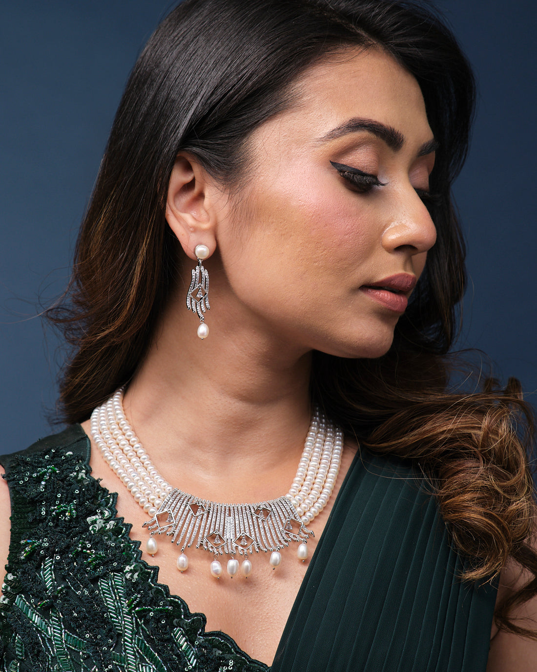 The Rang Tarangini Pearl Necklace Set by Chandrani Pearls, featuring teardrop and geometric details, is elegantly displayed with matching earrings on a green velvet stand.