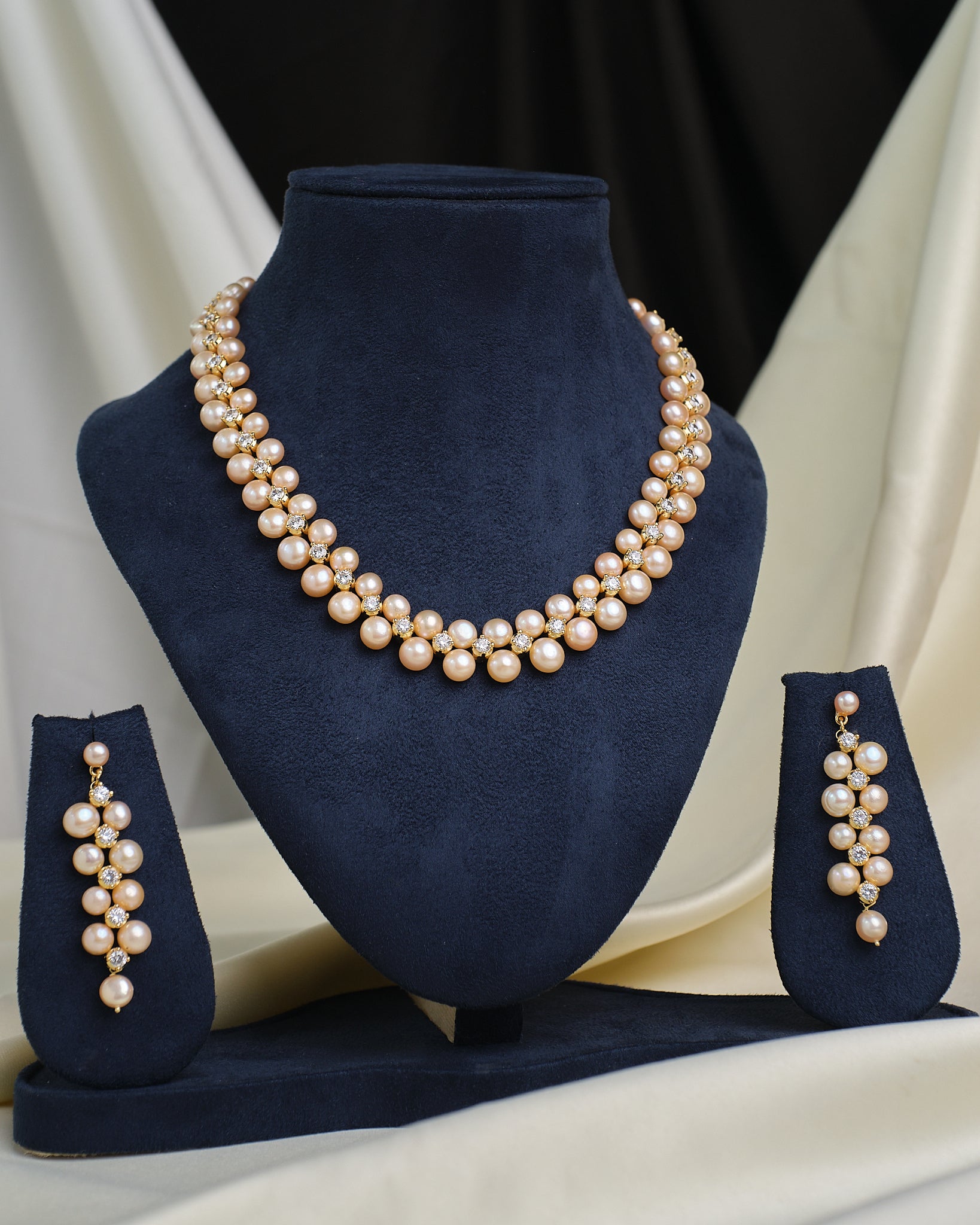 The Royal Mikkimotor Pink CZ Crown Set by Chandrani Pearls, featuring a pearl necklace with a pink CZ crown pendant and matching earrings, is displayed on navy blue velvet stands against a cream and black fabric background.