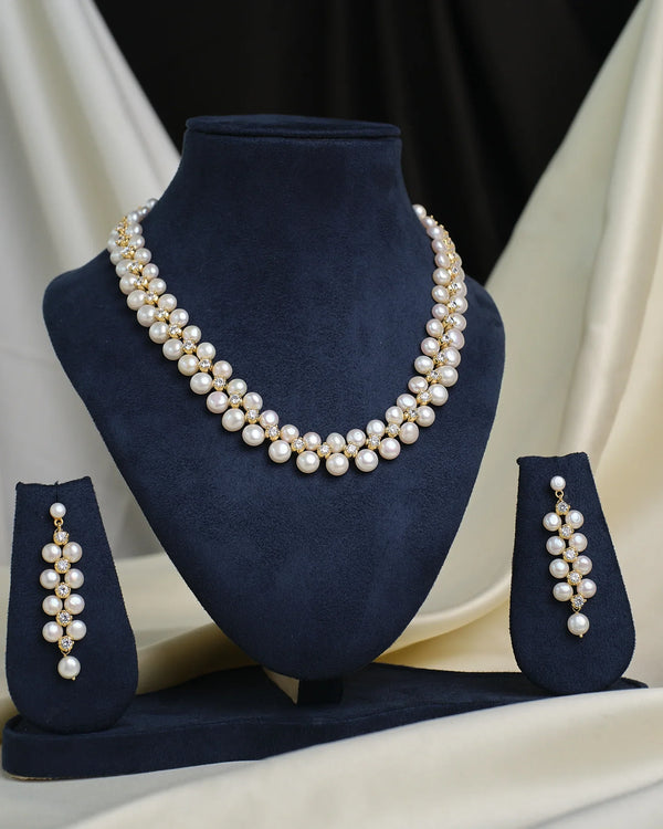 The Royal Mikkimotor White CZ Crown Set by Chandrani Pearls features a double-strand pearl necklace with gold accents and a crown-shaped pendant, displayed on a navy stand, paired with matching dangling pearl earrings.