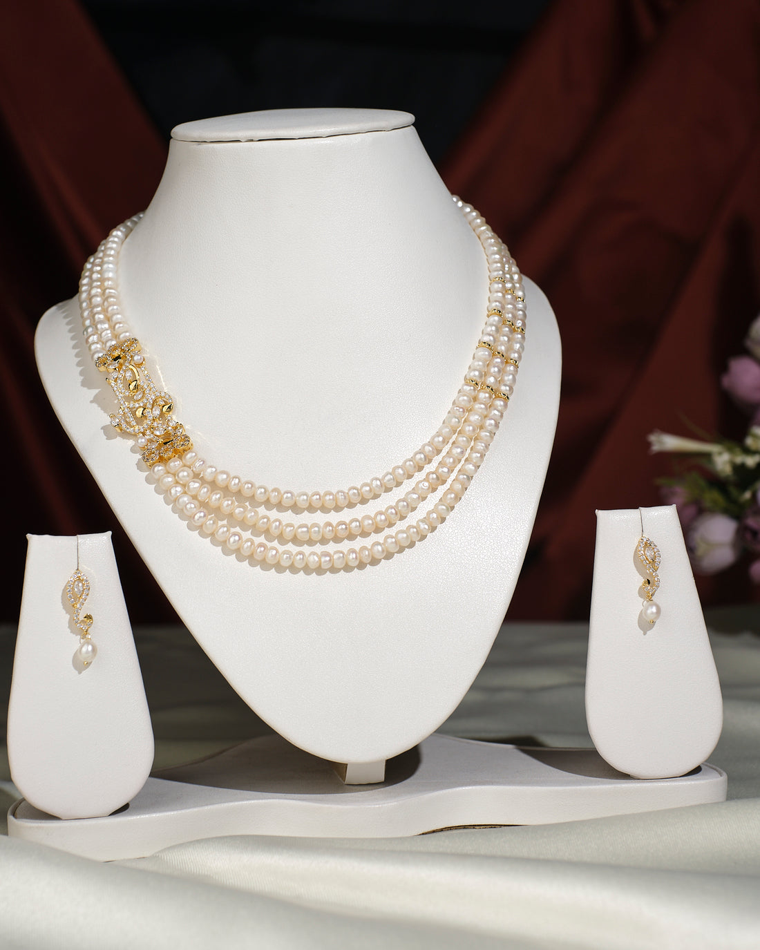 The Ravishing Pearl Necklace Set by Chandrani Pearls India is displayed on a white stand, featuring a gold clasp and matching pearl drop earrings on either side for a timeless, elegant look.