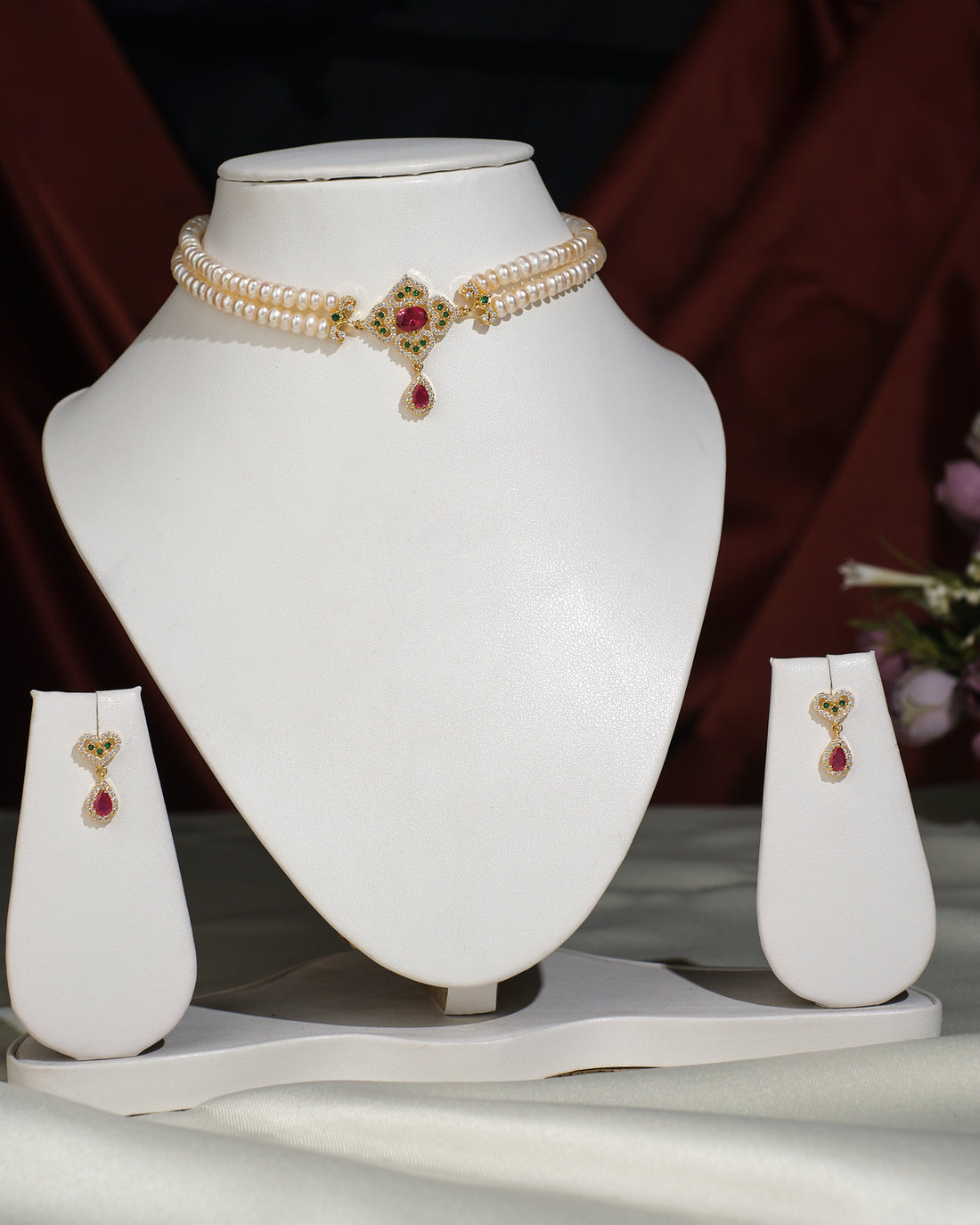 The Ravishing Pearl Choker Set by Chandrani Pearls India features a pearl choker with a central gold and red gemstone pendant, elegantly displayed on a white stand with matching earrings on separate stands for a coordinated look.