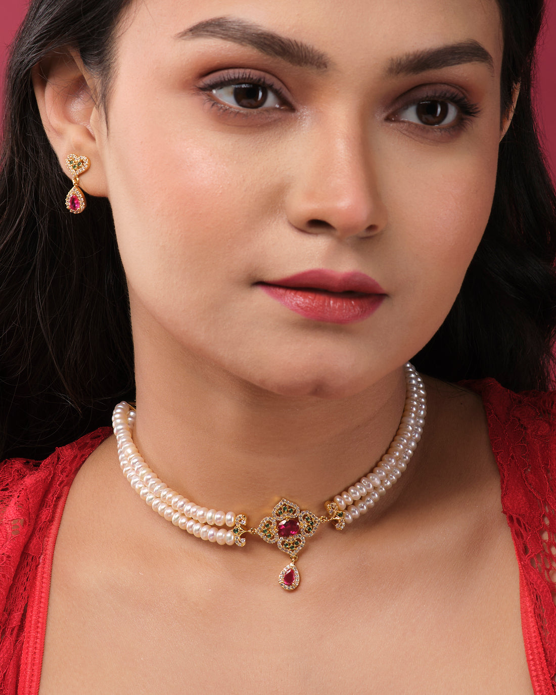 The Ravishing Pearl Choker Set by Chandrani Pearls India features a pearl choker with a central gold and red gemstone pendant, elegantly displayed on a white stand with matching earrings on separate stands for a coordinated look.