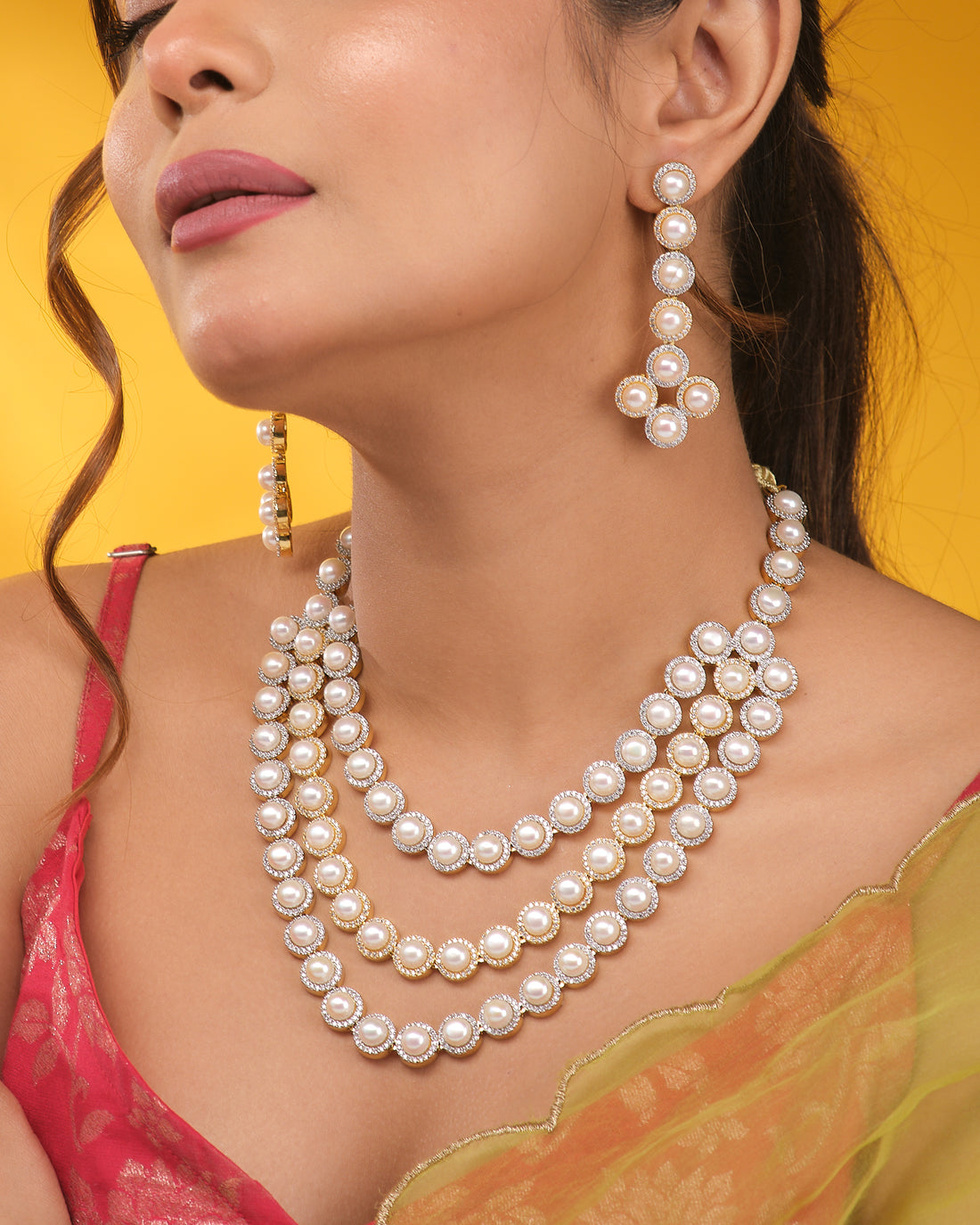 The Glamorous Glow CZ Necklace Set by Chandrani Pearls features a three-layered pearl and CZ necklace with a radiant center stone and matching pearl earrings, displayed on a navy blue stand against a cream and black fabric background.