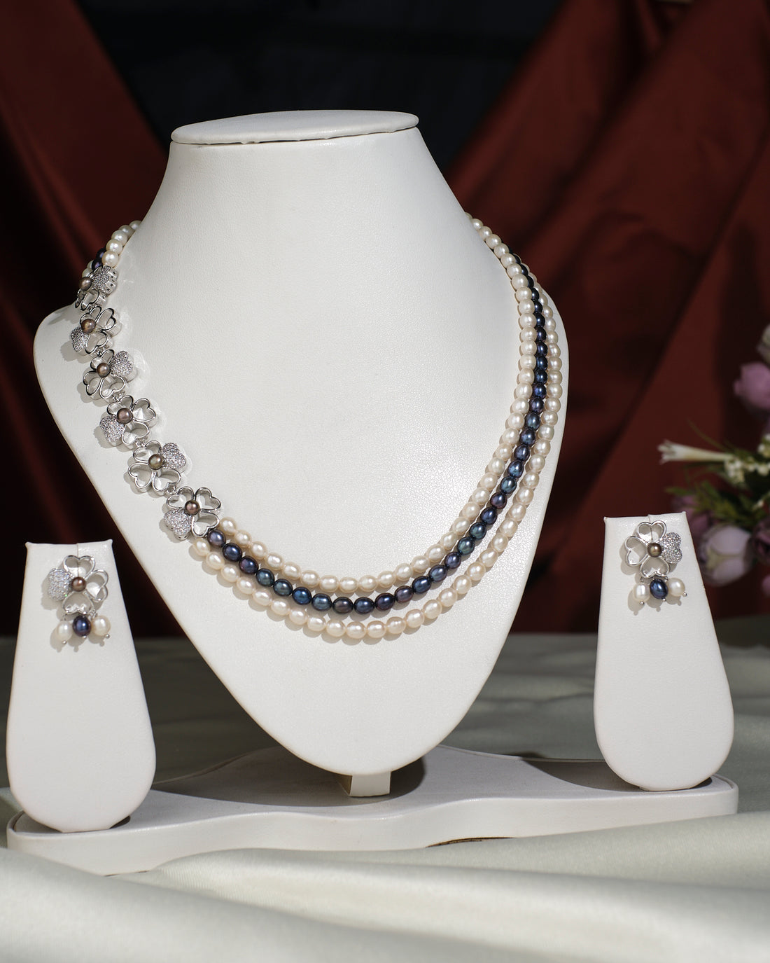 The Florious Side Pearl Necklace Set by Chandrani Pearls India features pearl and gemstone necklaces with floral earrings, elegantly displayed on a white stand against a light cloth background.