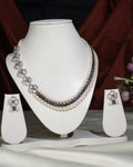 The Florious Side Pearl Necklace Set by Chandrani Pearls India features pearl and gemstone necklaces with floral earrings, elegantly displayed on a white stand against a light cloth background.