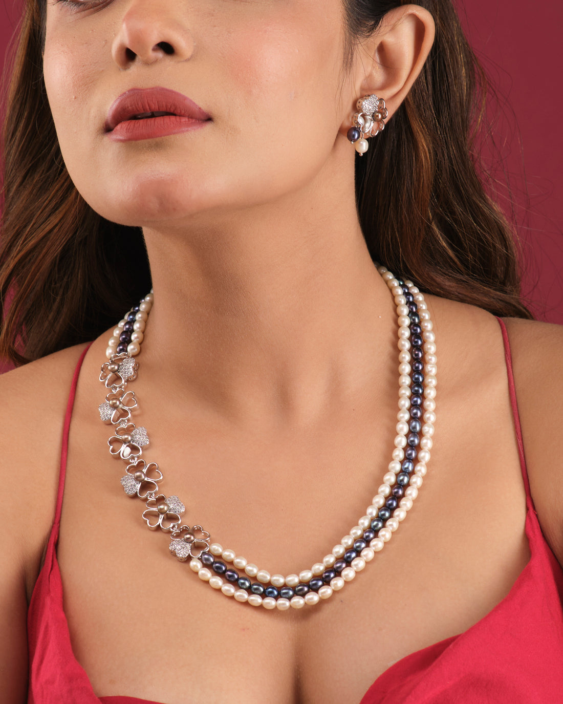 The Florious Side Pearl Necklace Set by Chandrani Pearls India features pearl and gemstone necklaces with floral earrings, elegantly displayed on a white stand against a light cloth background.