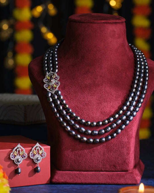 The Nakshatra Pearl Necklace Set by Chandrani Pearls, featuring four strands and a decorative brooch, is elegantly displayed on a red velvet bust, complemented by matching earrings on a red box.