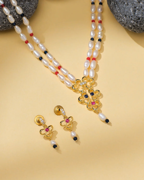 The Graceful Beads & Pearl Elegance Set by Chandrani Pearls features a gold and pearl necklace with red and black bead accents, plus matching earrings, exuding timeless sophistication on a beige surface with gray stones.