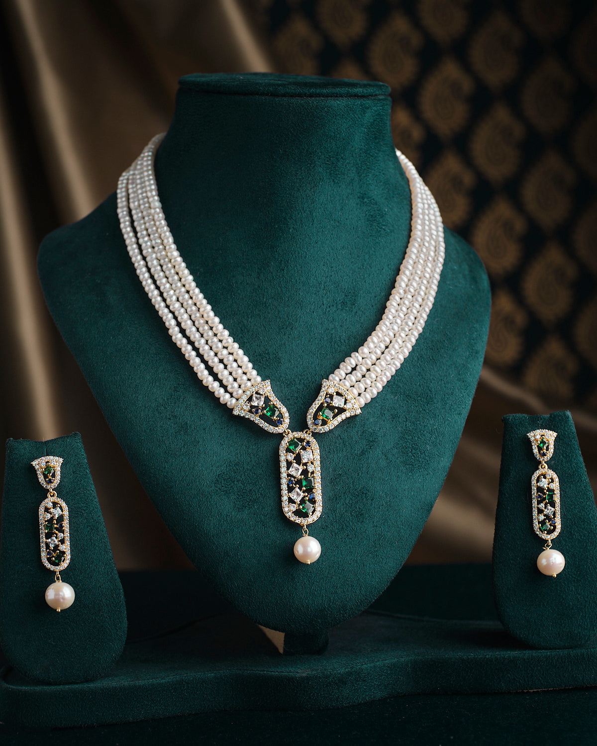 The Saratas - Mystic Mints Pearl Necklace Set by Chandrani Pearls, featuring a multi-strand design with pearls and emeralds, complete with matching earrings, is beautifully displayed on a dark green stand.