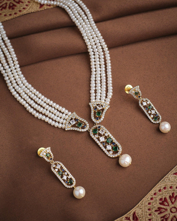 The Queen’s Aura Pearl CZ Necklace Set by Chandrani Pearls is a three-layer pearl necklace with matching earrings, adorned with gold, green, and white CZ stones for a royal look, shown on brown fabric with decorative borders.