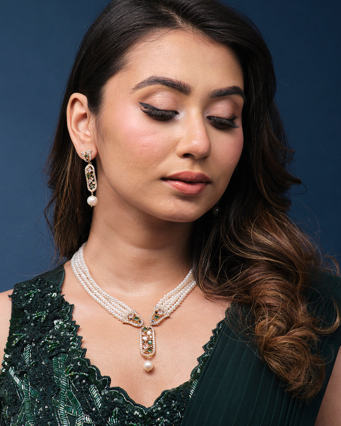 The Saratas - Mystic Mints Pearl Necklace Set by Chandrani Pearls, featuring a multi-strand design with pearls and emeralds, complete with matching earrings, is beautifully displayed on a dark green stand.