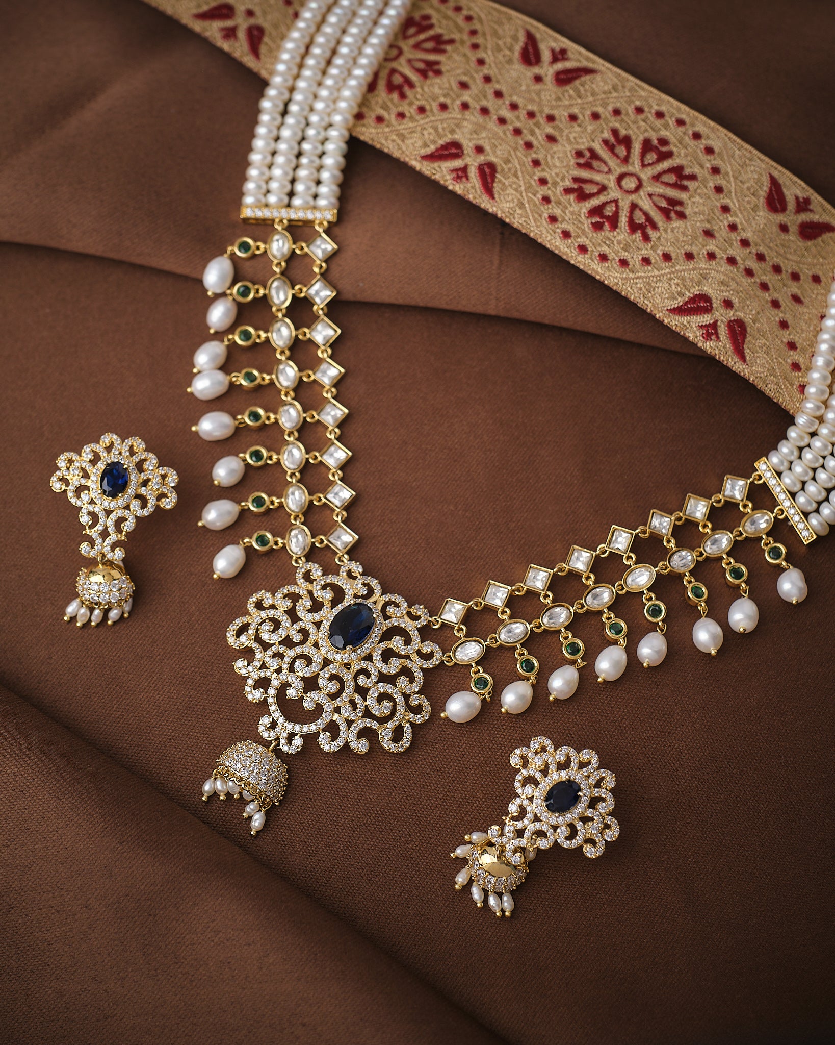 The Royal Celestial Pearl Light Necklace Set by Chandrani Pearls, featuring pearls, blue gemstones, CZ accents, and matching earrings, is displayed on brown fabric with a patterned textile in the background.