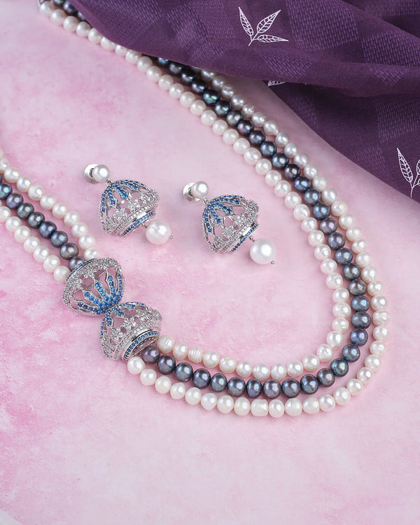 Chandrani Pearls' Side Pendant Necklace—Subtle Elegance with a Modern Twist—paired with blue-accented jhumka earrings, radiates style and sophistication on a pink surface next to folded purple fabric.