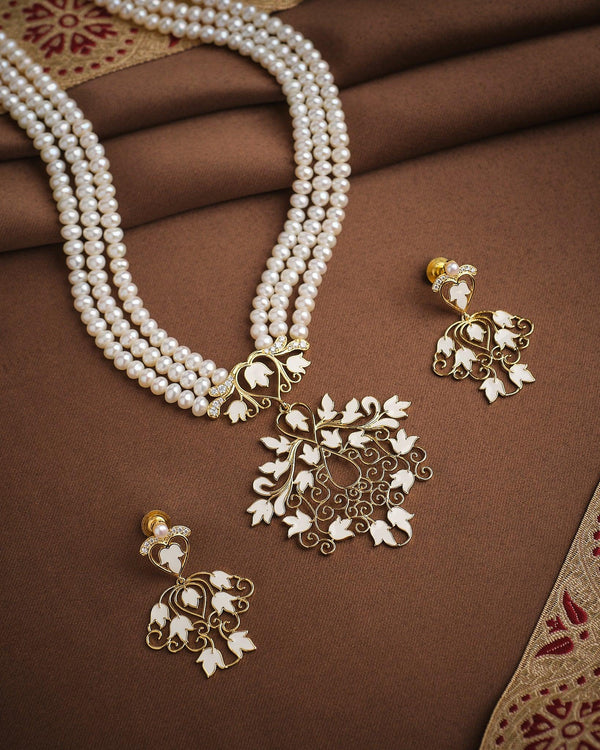 The Noor Majesty Pearl CZ Necklace Set by Chandrani Pearls features a pearls necklace with a gold and white floral pendant and matching earrings accented with CZ halos, elegantly displayed on brown fabric.