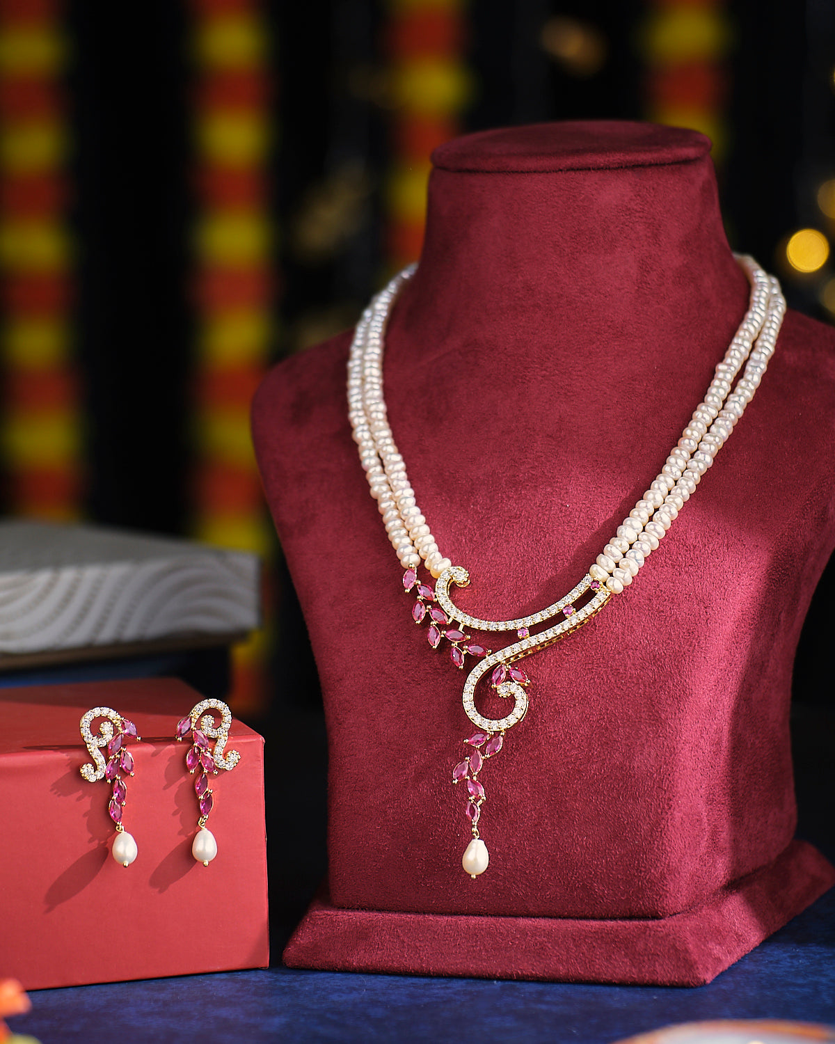 The Chaandikusuma - The Royal Bloosom Pearl Necklace Set by Chandrani Pearls features intricate gold and pink accents and is elegantly presented on a red velvet stand, alongside matching earrings in a red box.