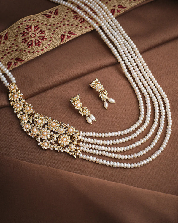 The Flory Pearl & CZ Necklace Set by Chandrani Pearls features a multi-strand pearl necklace with gold floral details and matching earrings, displayed on brown fabric beside a decorative gold and red textile.