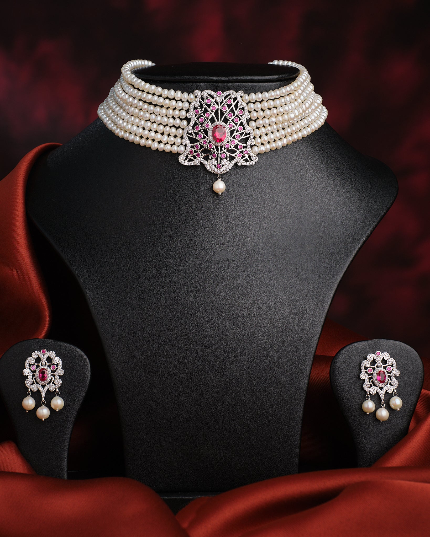 The Somalekha-Modern Yet Traditional Pearl Necklace Set by Chandrani Pearls features a central silver and pink floral pendant with matching earrings, elegantly displayed on a black stand against red satin fabric.