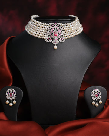 The Somalekha-Modern Yet Traditional Pearl Necklace Set by Chandrani Pearls features a central silver and pink floral pendant with matching earrings, elegantly displayed on a black stand against red satin fabric.
