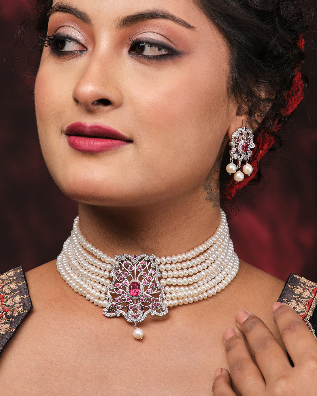 The Somalekha-Modern Yet Traditional Pearl Necklace Set by Chandrani Pearls features a central silver and pink floral pendant with matching earrings, elegantly displayed on a black stand against red satin fabric.