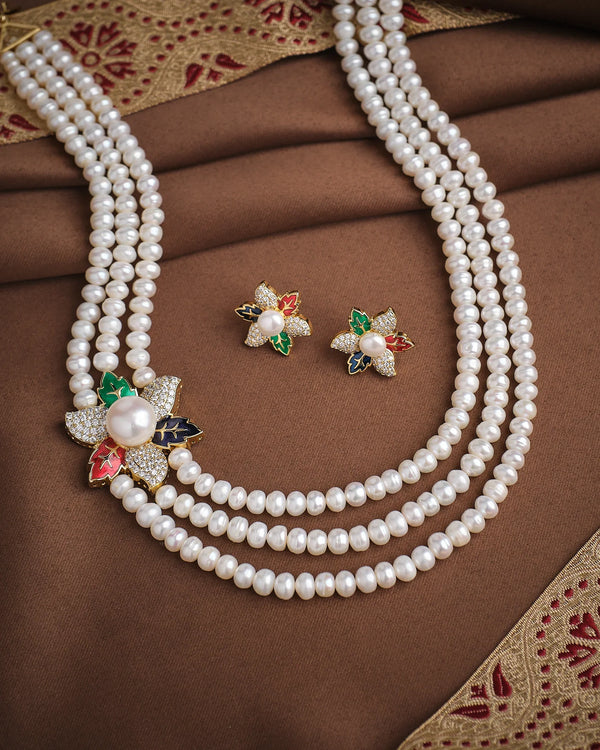 The Royal Floral Pearl Necklace Set by Chandrani Pearls features three layers of pearls, floral gemstone accents, a tri-color pendant, sparkling CZ stones, and comes with matching earrings, elegantly displayed on brown embroidered fabric.