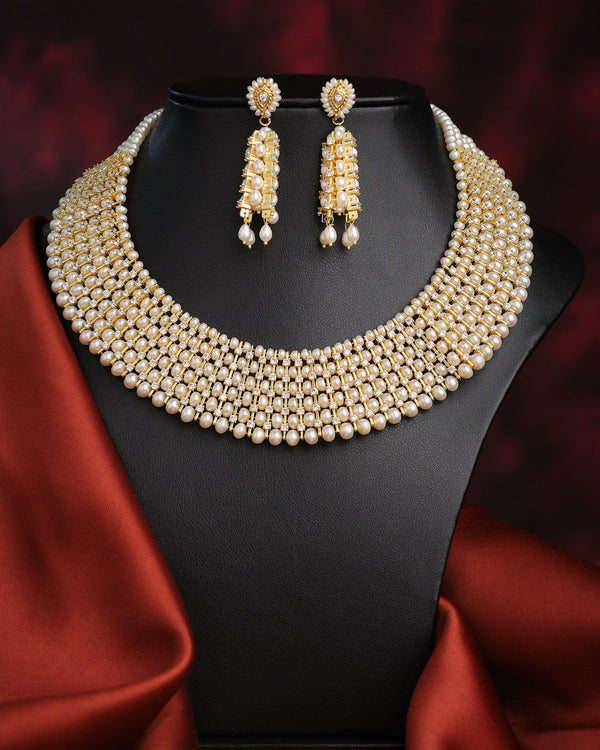 The Suvarnachandra-Golden Moon Royal Pearl Necklace Set by Chandrani Pearls, featuring gold and pearl with crystal accents and matching earrings, is displayed on a black jewelry stand draped with satin red fabric.