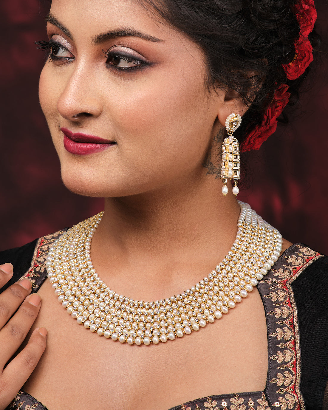 The Suvarnachandra-Golden Moon Royal Pearl Necklace Set by Chandrani Pearls, featuring gold and pearl with crystal accents and matching earrings, is displayed on a black jewelry stand draped with satin red fabric.