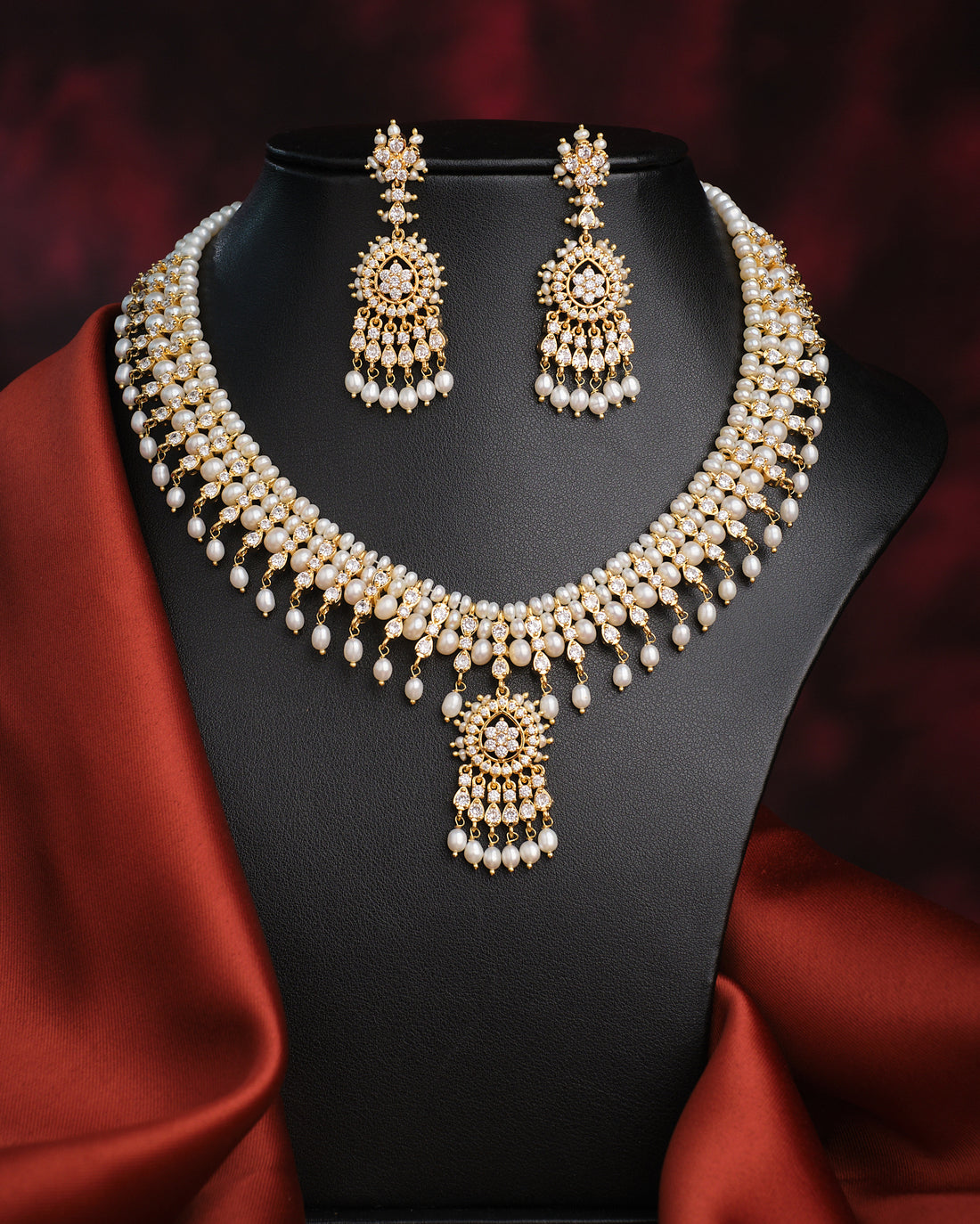 The Suryaprabha-Luxe Heritage Pearl Necklace Set by Chandrani Pearls, displayed with matching earrings on a black bust against a dark backdrop and red fabric accent, exudes timeless elegance.
