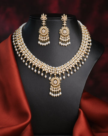 The Suryaprabha-Luxe Heritage Pearl Necklace Set by Chandrani Pearls, displayed with matching earrings on a black bust against a dark backdrop and red fabric accent, exudes timeless elegance.