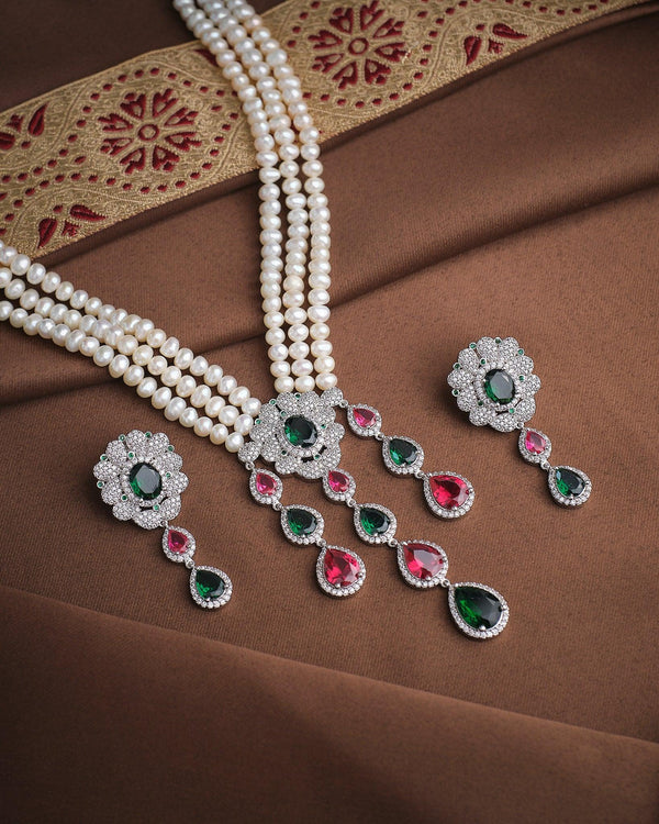 The Gorgeous Dazzle Pearl CZ Set by Chandrani Pearls, featuring luminous pearls with diamond, green, and pink gemstone pendants and matching earrings, is displayed on brown fabric with a gold and red embroidered border.