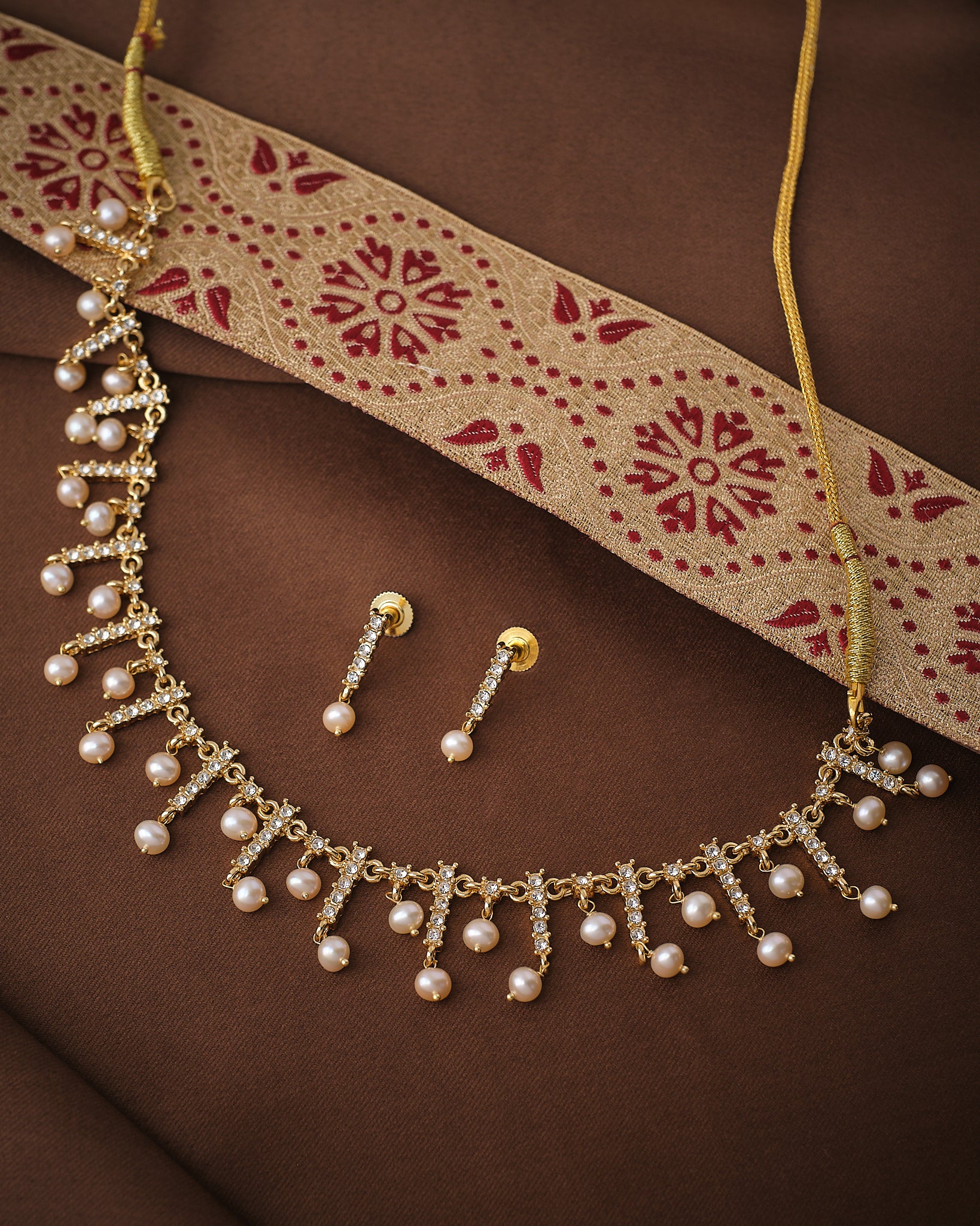 The Pearl & CZ Imperial Layers Necklace Set by Chandrani Pearls, featuring pearl drops, CZ motifs, and matching gold and pearl earrings, is displayed on brown fabric with a gold and red embroidered cloth.