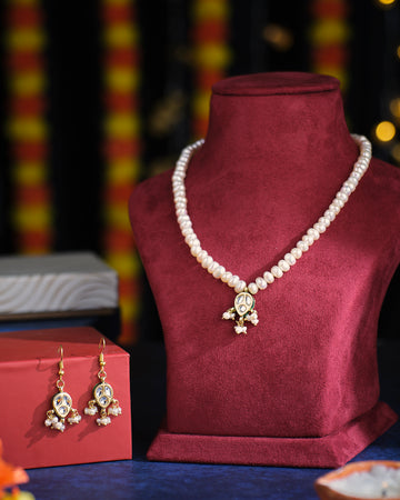 The Keyair Pearl Necklace Set by Chandrani Pearls, featuring a dangling pendant, is elegantly displayed on a red velvet stand and comes with matching earrings presented in a box.