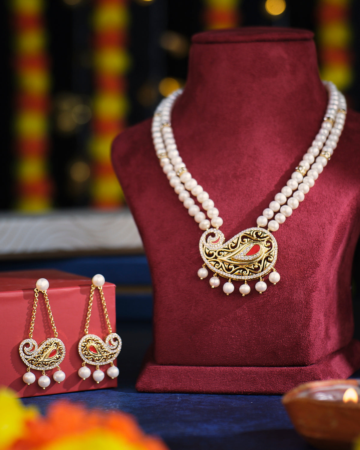 The Fallen Swarnadhara Pearl Necklace Set by Chandrani Pearls is showcased on a burgundy stand, featuring a gold and red pendant. Matching earrings lie close by, positioned beside a small red box.