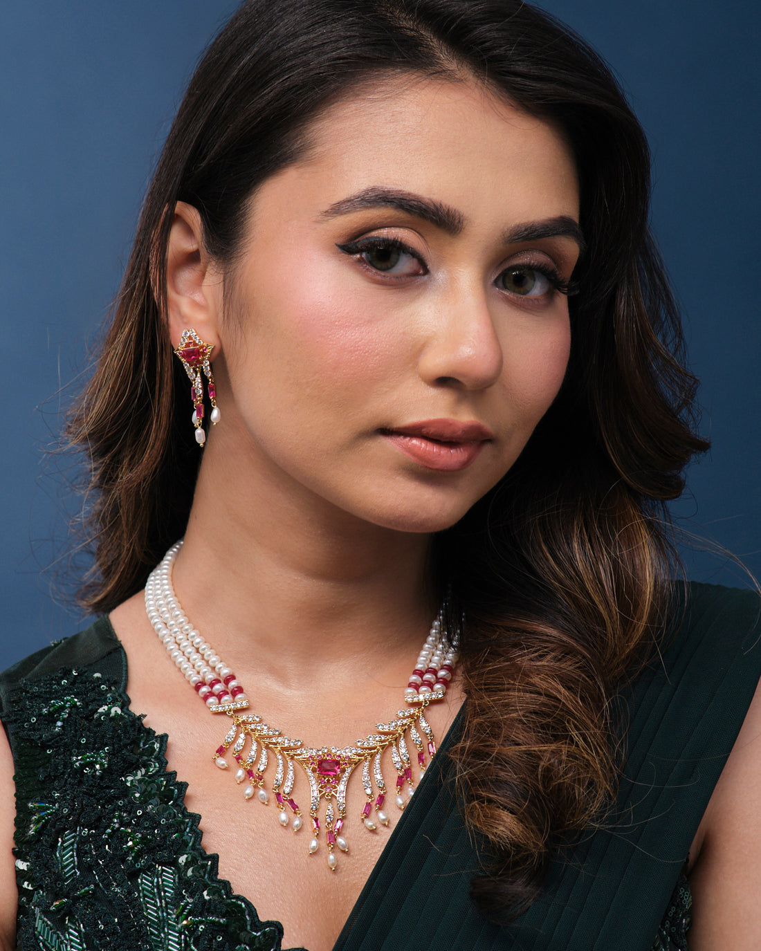 The Anvesha Necklace Set by Chandrani Pearls India, featuring a pearl and gemstone necklace with matching earrings, is showcased on a dark green stand.