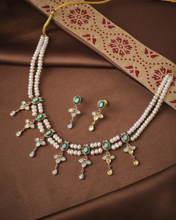 The Queen’s Radiant Pearl CZ Set by Chandrani Pearls features a pearl and gold necklace with green and white CZ stones, matching earrings, and a brown and gold embroidered cloth displayed on a brown background.