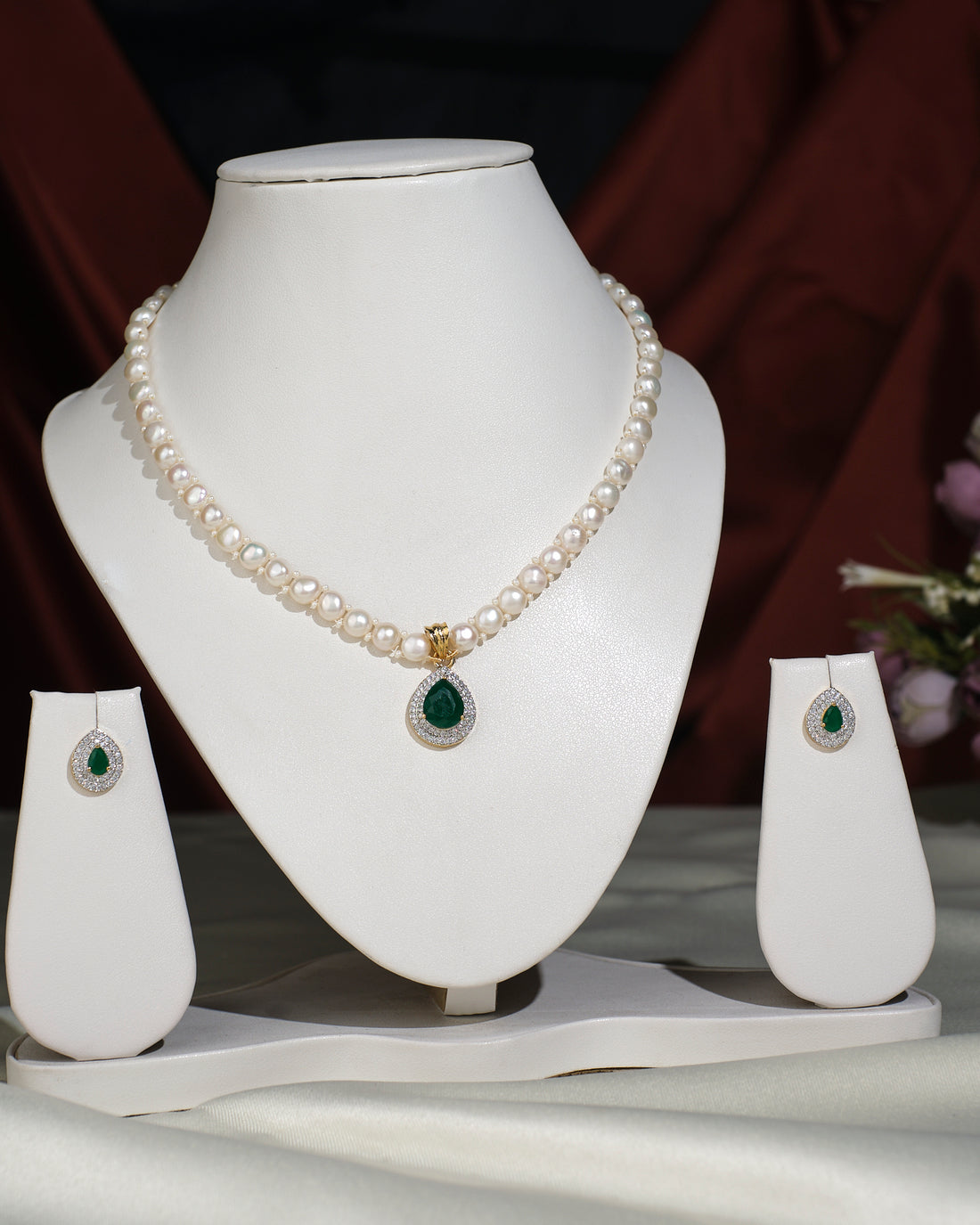 The Elegant Pearl Necklace Set by Chandrani Pearls India, featuring matching earrings with green gemstone pendants, is showcased on white stands against a bold two-tone background, ready to enhance any ensemble.