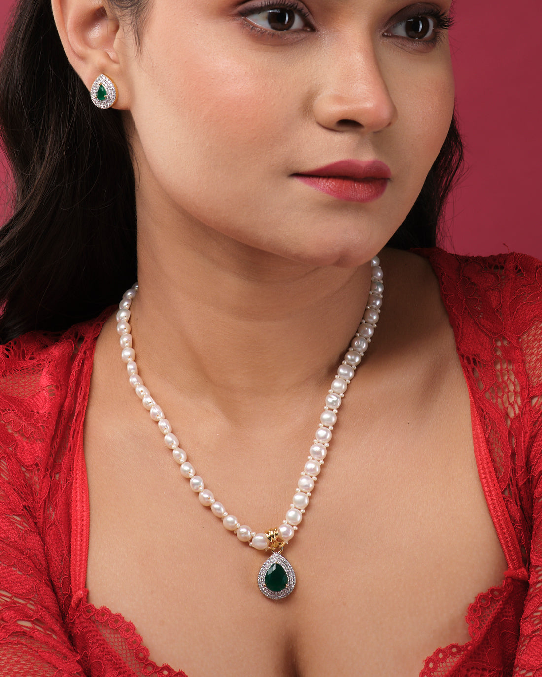 The Elegant Pearl Necklace Set by Chandrani Pearls India, featuring matching earrings with green gemstone pendants, is showcased on white stands against a bold two-tone background, ready to enhance any ensemble.