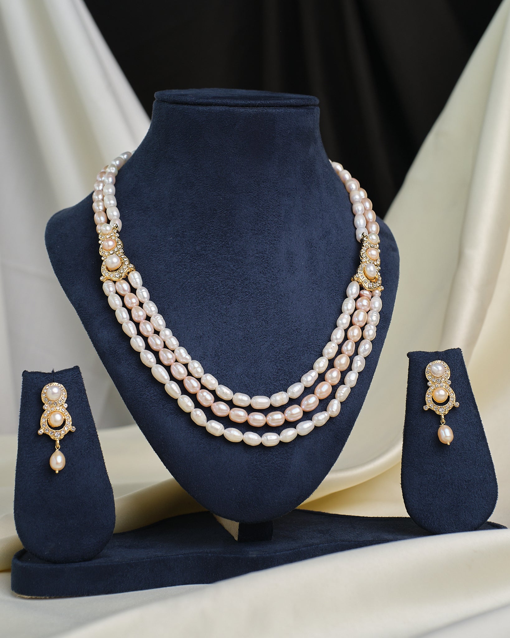 The Chandrani Pearls Layered CZ Necklace & Earring Set is showcased on navy blue velvet stands, creating an elegant and dramatic effect against a draped fabric background.