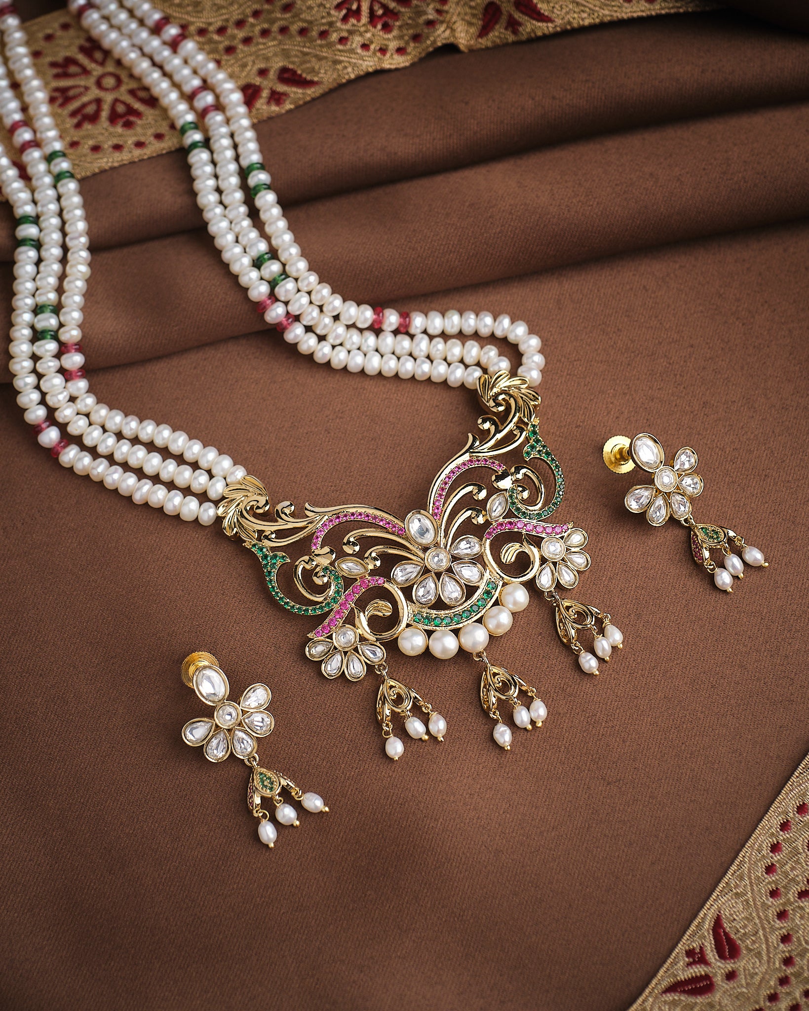 The Noor Royal Embrace Necklace Set by Chandrani Pearls features traditional pearl and gold intricate designs, CZ bezels, and matching earrings, elegantly displayed on brown fabric.