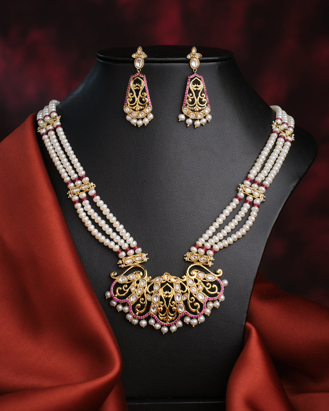 The Rangamaya-Filled With Colours Pearl Necklace Set by Chandrani Pearls, featuring gold, white, and pink beads with intricate design and matching earrings, is showcased on a black bust draped in red satin.
