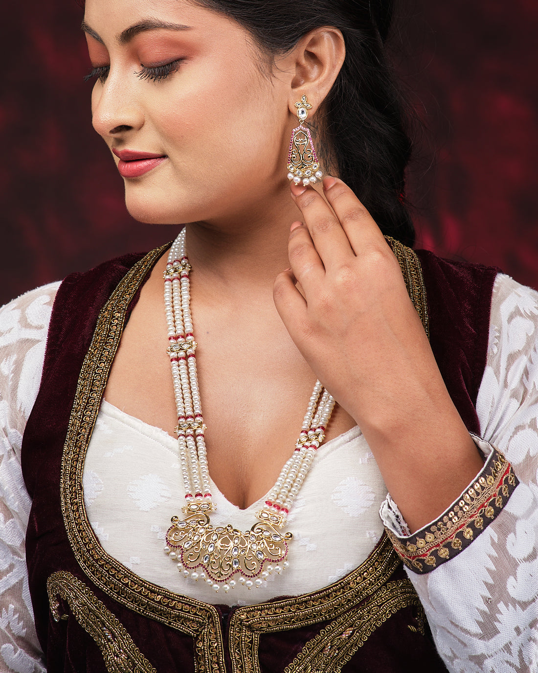 The Rangamaya-Filled With Colours Pearl Necklace Set by Chandrani Pearls, featuring gold, white, and pink beads with intricate design and matching earrings, is showcased on a black bust draped in red satin.
