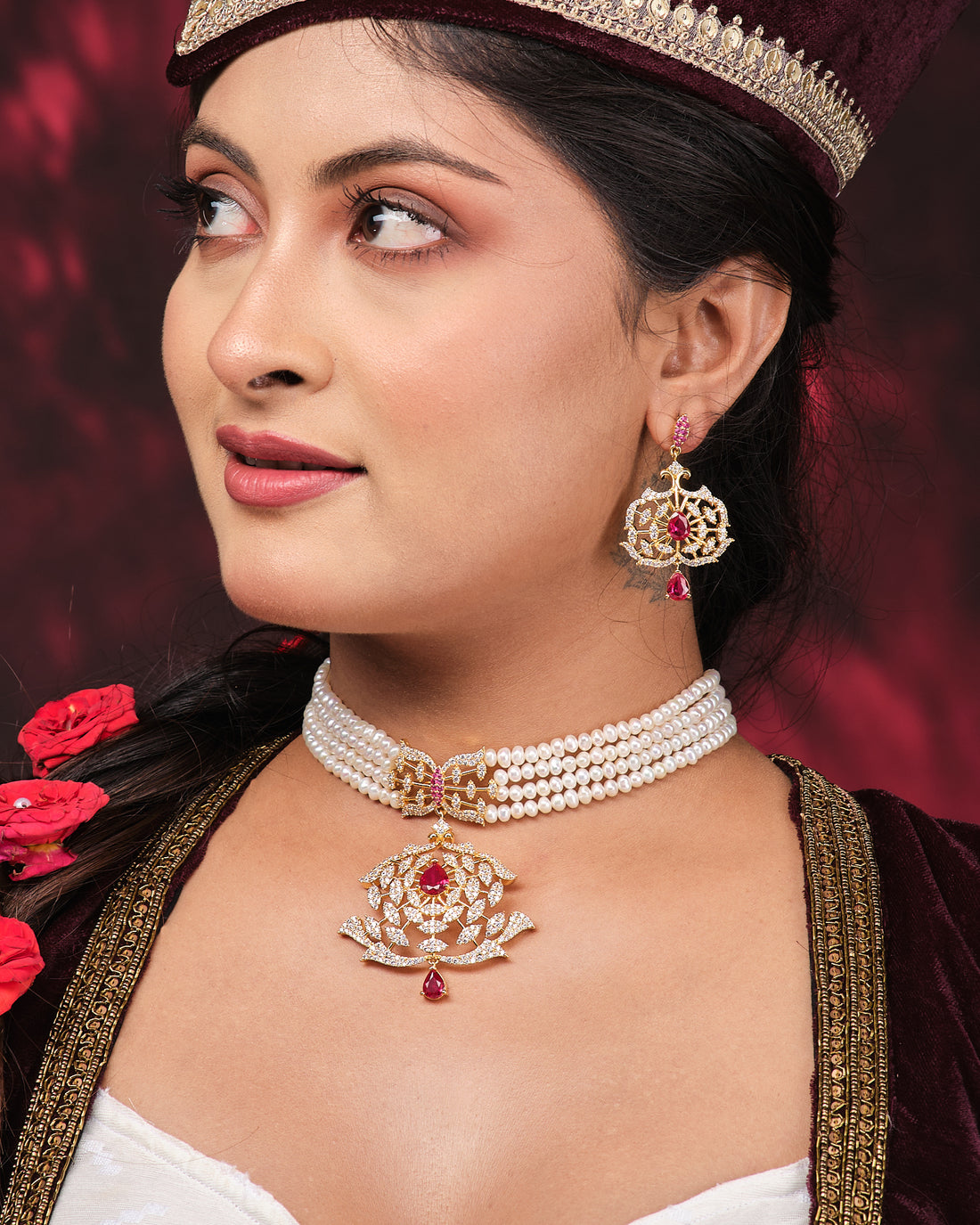 The Tribandhura Pearl Necklace Set by Chandrani Pearls, featuring a gold and red gemstone pendant on a pearl choker with matching earrings, is displayed on a black stand against a dark red draped background.