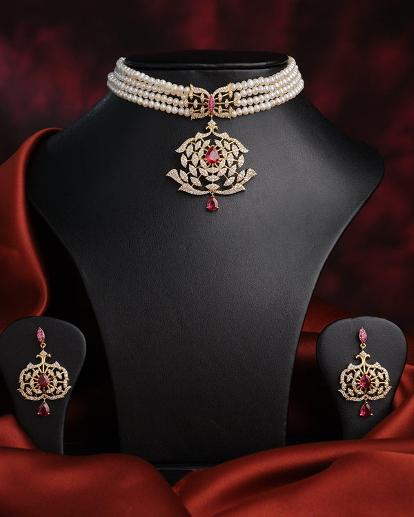The Tribandhura Pearl Necklace Set by Chandrani Pearls, featuring a gold and red gemstone pendant on a pearl choker with matching earrings, is displayed on a black stand against a dark red draped background.