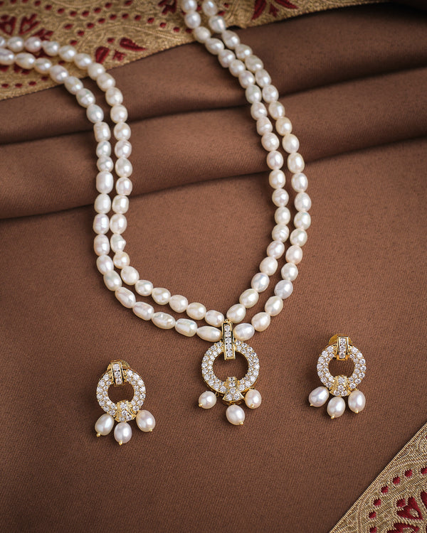 The Pretty CZ Pearl Necklace Set by Chandrani Pearls features radiant double-strand pearls with a gold and diamond pendant, paired with matching earrings, elegantly displayed on brown and maroon fabric.