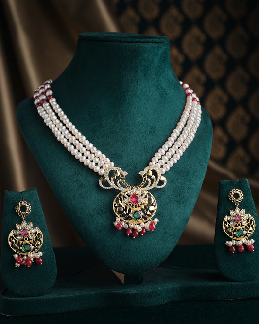 The Rajeshwari Pearl Necklace Set by Chandrani Pearls includes elegant pearl necklace and earrings adorned with intricate gold embellishments and red and green gemstones, displayed on a green velvet stand against a patterned background.