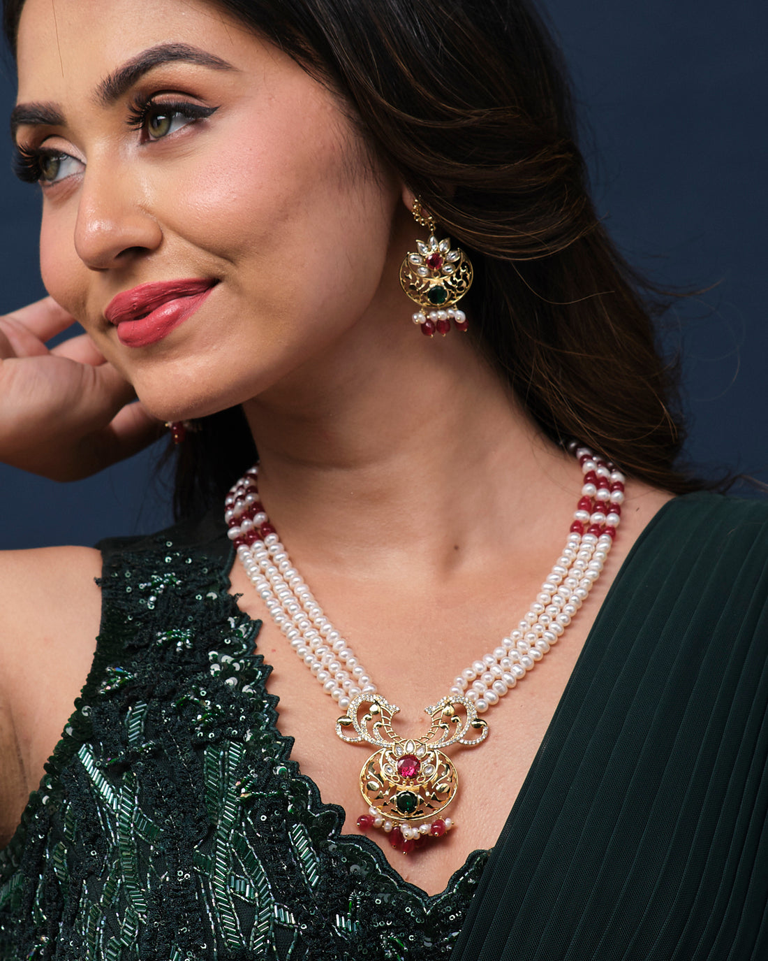 The Rajeshwari Pearl Necklace Set by Chandrani Pearls includes elegant pearl necklace and earrings adorned with intricate gold embellishments and red and green gemstones, displayed on a green velvet stand against a patterned background.