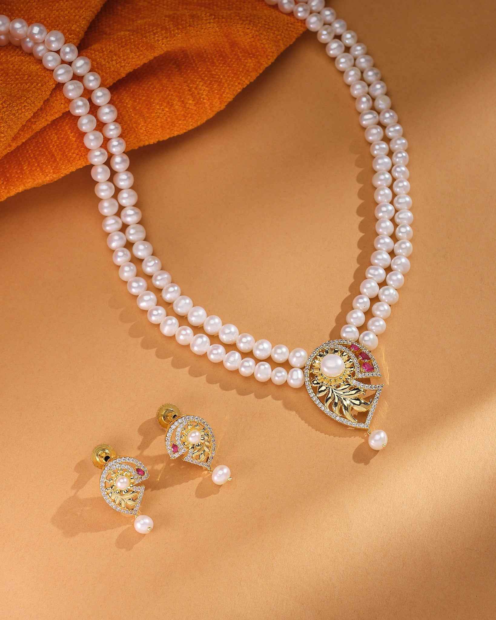 The Celina CZ Pearl Set by Chandrani Pearls—featuring a refined double-strand pearl necklace with a gold and gemstone pendant plus matching pearl and gold earrings—radiates timeless brilliance on a tan background.