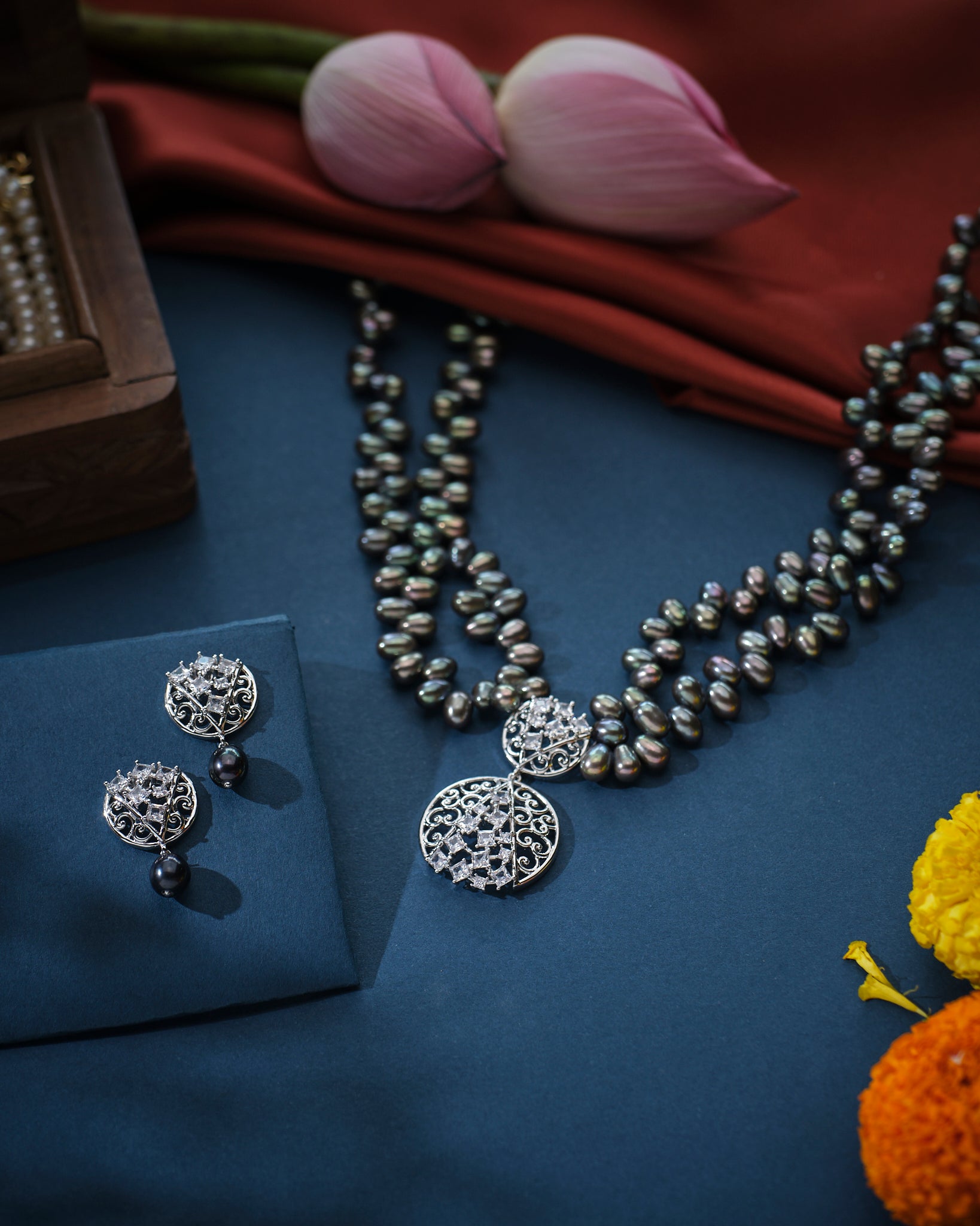 The "Out Of The Dark - Black Multilayered Pearl Necklace Set" by Chandrani Pearls, featuring an intricate round pendant and matching earrings, is elegantly showcased on a red velvet bust against a softly blurred festive backdrop adorned with lights and flowers.