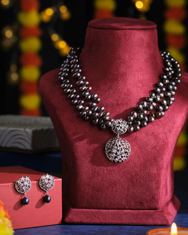 The "Out Of The Dark - Black Multilayered Pearl Necklace Set" by Chandrani Pearls, featuring an intricate round pendant and matching earrings, is elegantly showcased on a red velvet bust against a softly blurred festive backdrop adorned with lights and flowers.
