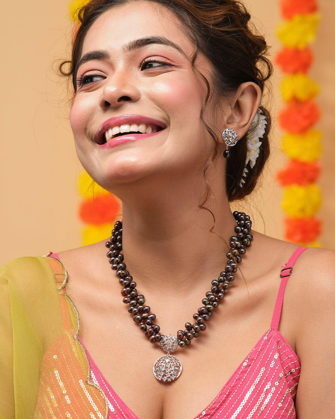 The "Out Of The Dark - Black Multilayered Pearl Necklace Set" by Chandrani Pearls, featuring an intricate round pendant and matching earrings, is elegantly showcased on a red velvet bust against a softly blurred festive backdrop adorned with lights and flowers.