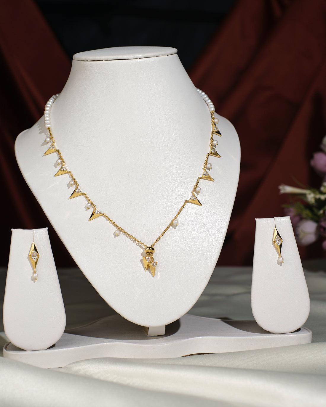 The Cute Necklace Set by Chandrani Pearls India features gold triangular pendants with pearl accents, displayed on a white stand with matching earrings, highlighting its natural beauty against draped fabric.